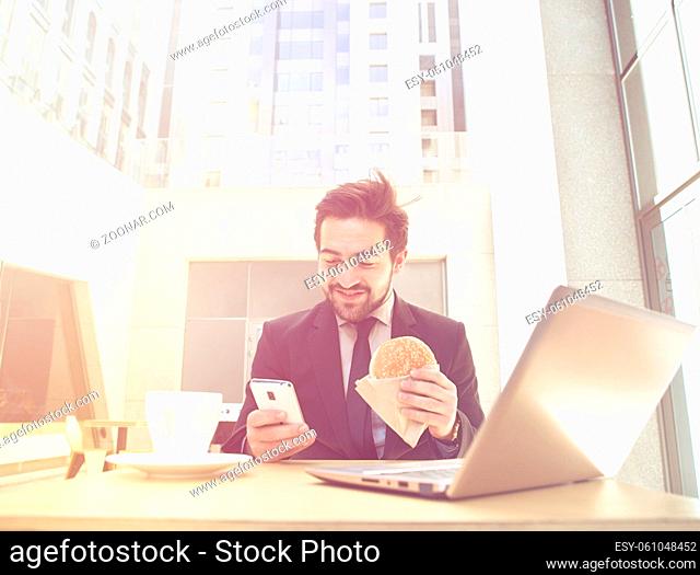 Toned picture of handsome businessman in black business suit using mobile phone while eating hamburger. Laptop computer is in front of him