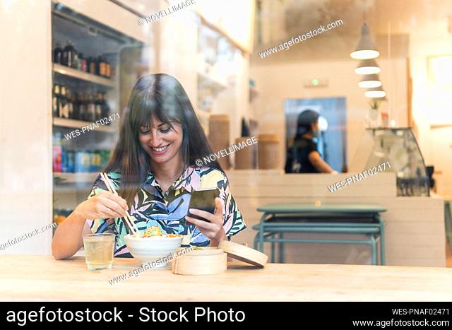 Smiling woman with smart phone having food at restaurant seen through glass