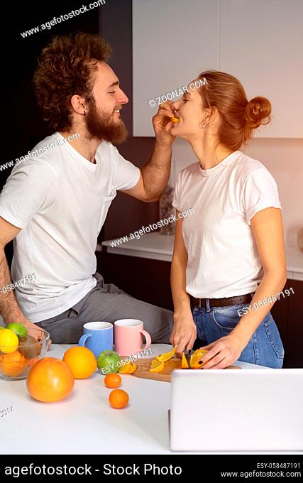 Feeding orange to girl young man embracing each other enjoying their their morning coffee in a new house. Making fun at modern kitchen and kissing while cooking...