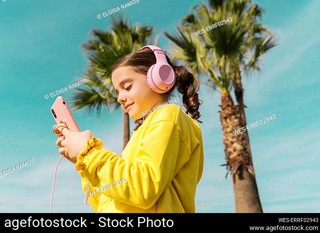 Portrait of smiling little girl wearing yellow jacket listening music with headphones looking at smartphone