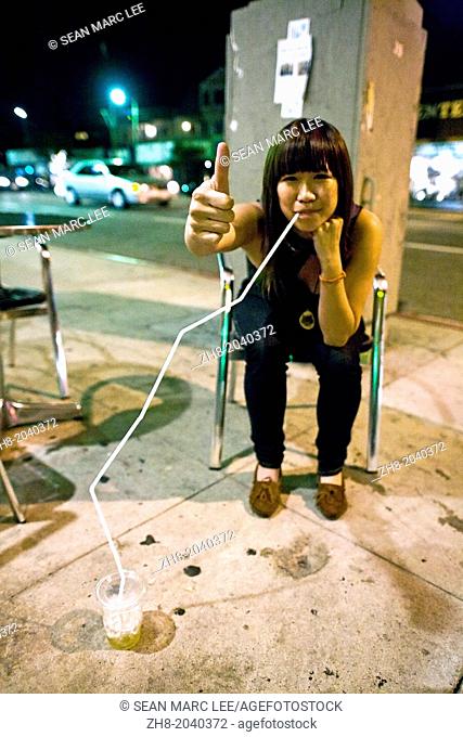A young Asian woman drinking from a very large straw while holding a thumbs up on the street in Silverlake, Los Angeles California
