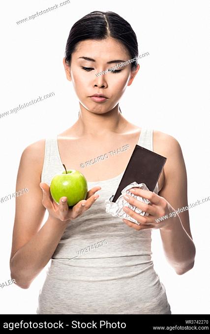 Pretty Asian woman debating an apple or chocolate as she stands with a fresh green apple in one hand and unwrapped bar of candy in the other in a healthy diet...