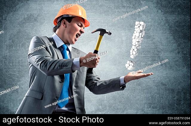 Furious businessman going to crash with hammer exclamation mark. Young handsome man in business suit and safety helmet standing on wall background
