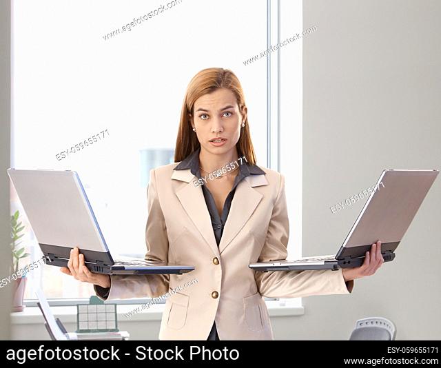 Desperate businesswoman standing in bright office holding two laptops in hands