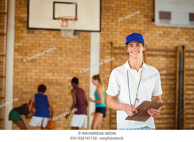 Coach smiling at camera while high school team playing basketball in background