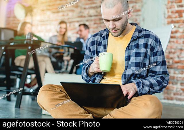 Hip worker in a startup coding with laptop sitting on skateboard with colleagues in background