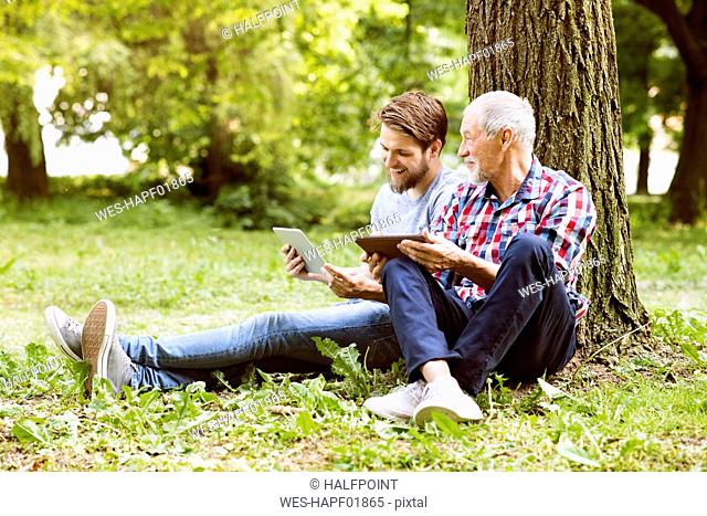 Senior father sitting besides his adult son in a park with tablet