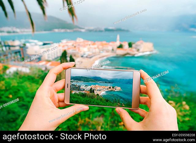 Men hold a smartphone with a photo of architecture in their hands. In the background is the old town of Budva, Montenegro. High quality photo