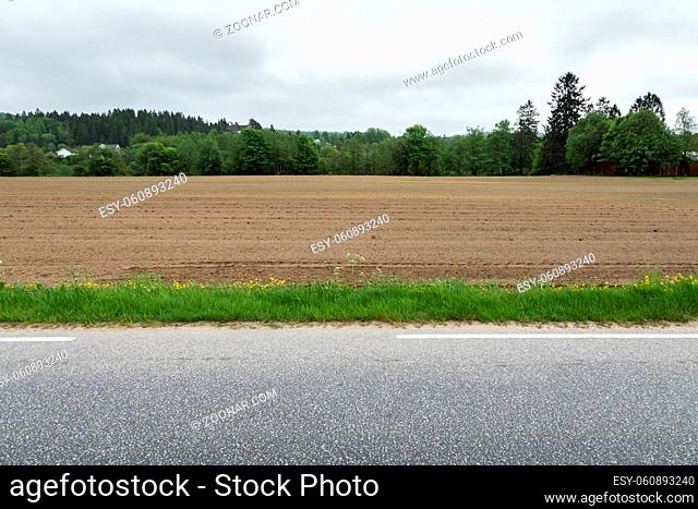 A brown field with ploughed rows of dirt next to a stripe of grass and an asphalt road on the countryside in Kristiansand, Norway