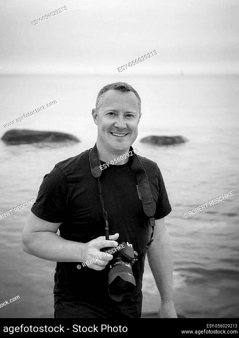 a male smiling photographer on tour on the beach to search for motives with a camera around his neck and in his hand in a black polo shirt with a view into the...