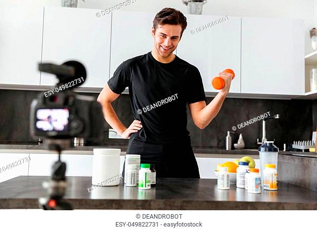 Happy young man filming his video blog episode about healthy food additives while standing at the kitchen table and exercising with a dumbbell