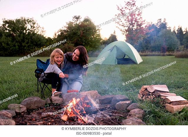 Mother and daughter sitting beside campfire, cooking sausage over fire