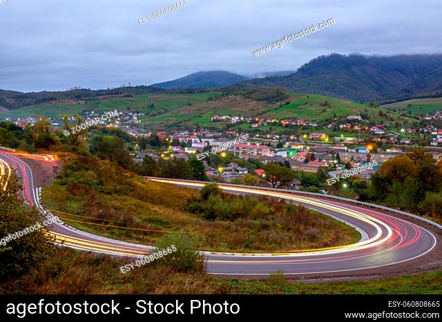 Cloudy evening on a mountain road. Headlight trails of fast moving cars. A small village shines with lights in the valley