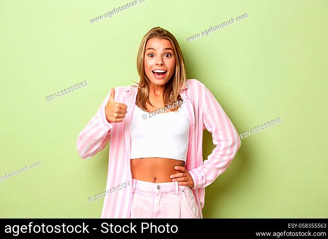 Image of surprised happy female model with blond hair, wearing pink shirt and jeans, showing thumbs-up in approval, like something