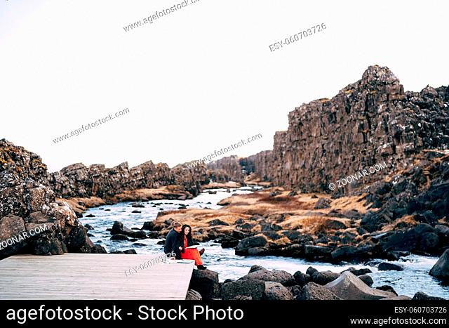 A guy and a girl sit hugging on a bridge near Ehsaraurfoss Falls, Ehsarau River, National Park, Tingvedlir, Sudurland Region, Iceland