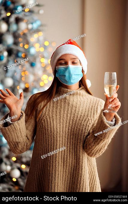 Beautiful young woman in protective mask with glass of champagne at home. Christmas celebration. The concept of celebrating new year and Christmas under...