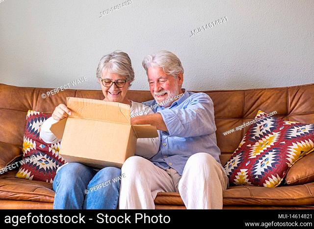Old couple unpacking delivery box at home. Happy senior couple looking at carton box while sitting on sofa in living room at home