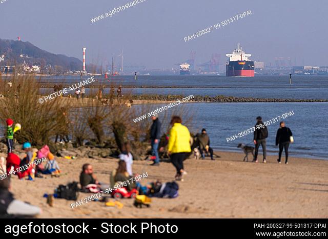 27 March 2020, Hamburg: People are sitting or walking on the Elbe beach in Wittenbergen while in the background a tanker and a container ship are sailing on the...