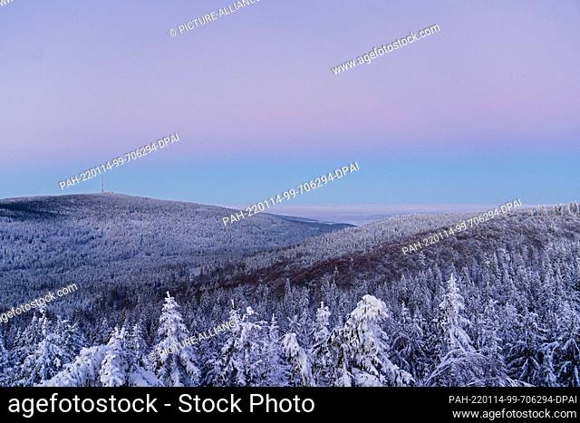 14 January 2022, Bavaria, Neubau: The sky above the snow-covered trees on the Nußhardt in the Fichtelgebirge is bathed in pastel colors by the dawn