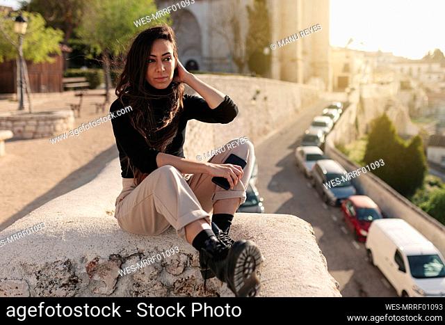 Tourist with mobile phone sitting on retaining wall during sunny day