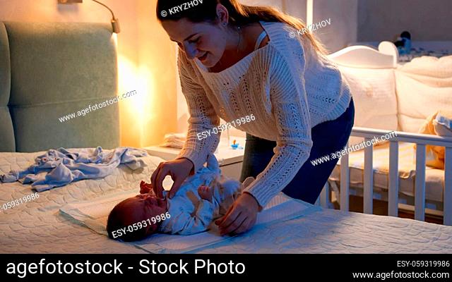 Beautiful smiling mother holding and cuddling her little newborn baby boy at night in bedroom. Mother changes clothes to little child