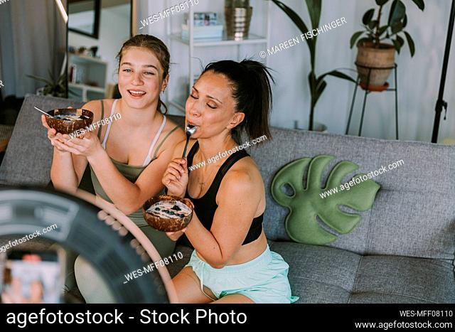 Female fitness trainers eating fruit bowls while vlogging at home
