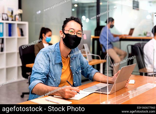 Portrait of asian office employee businessman wear protective face mask work in new normal office with interracial colleague in background as social distance...