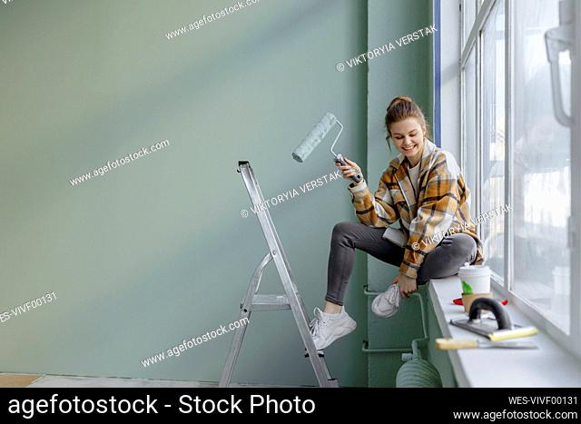 Smiling woman with paint roller sitting on window sill at home