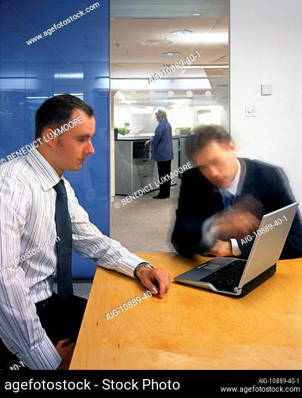 Office life and interiors. Two businessmen sitting at meeting table and woman by filing cabinets in background