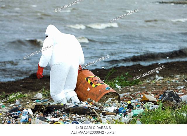 Person in protective suit carrying barrel of hazardous waste on polluted shore