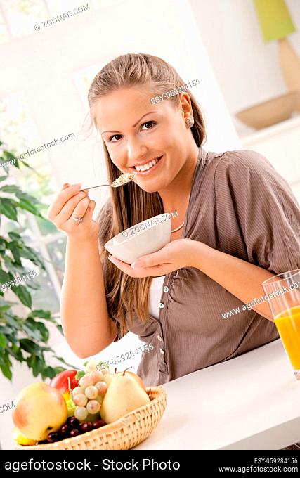 Happy young woman eating breakfast cereal and drinking orange juice, smiling