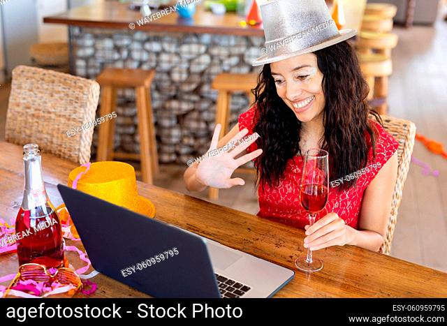 Smiling biracial young woman waving hand while enjoying party on video call over laptop at cafe