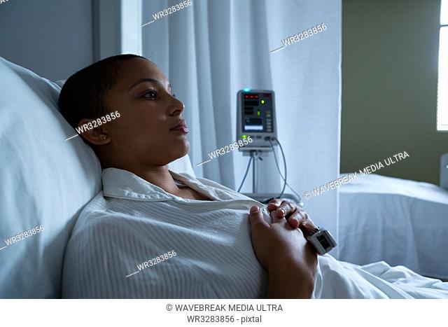 Side view of thoughtful mixed race female patient lying in bed with hands on her breasts in the ward in hospital. Empty bed and monitor are visible in the...