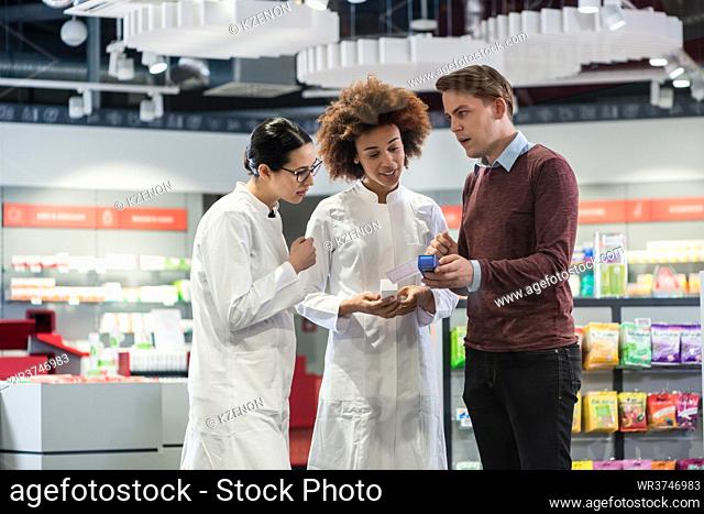 Young man asking for advice and opinion from two experienced female pharmacists regarding a prescribed pharmaceutical product in the interior of a modern...