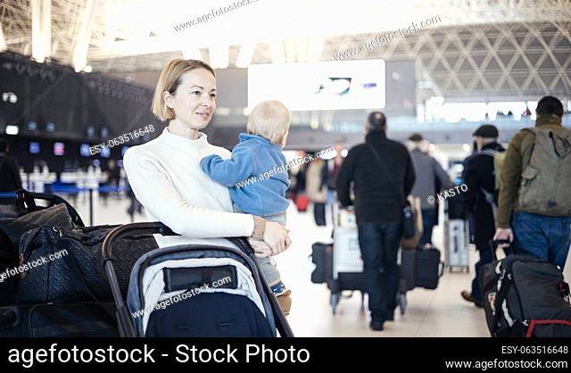 Motherat travelling with his infant baby boy child, walking, pushing baby stroller and luggage cart at airport terminal station