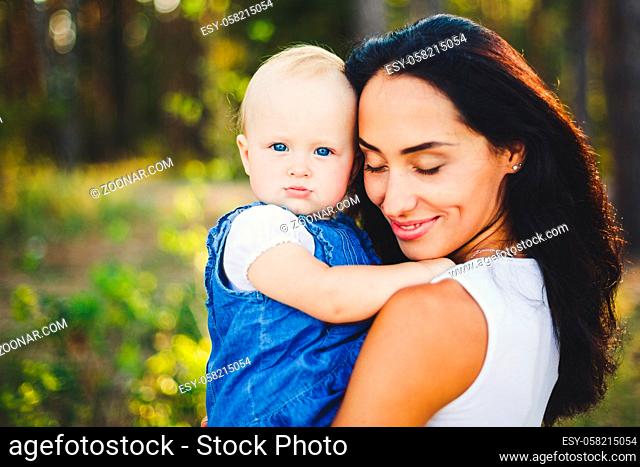 young beautiful fashionable mother with long brunette hair holding a daughter's blonde with blue eyes one year of birth in a coniferous forest in a summer park