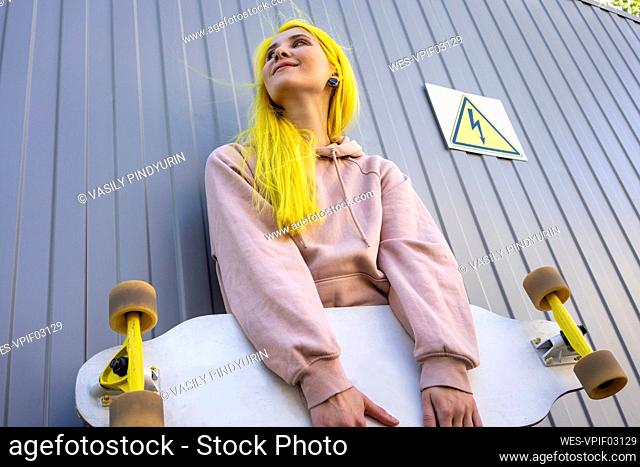 Young woman looking away holding skateboard standing against metal wall