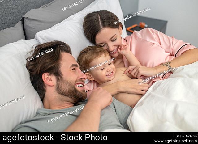 Happy people. Young smiling woman hugging touching cute baby and man lying together under blanket in bedroom at home