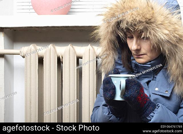 Middle age female model in parka and mittens hold coffee cup sitting on the floor under windowsill