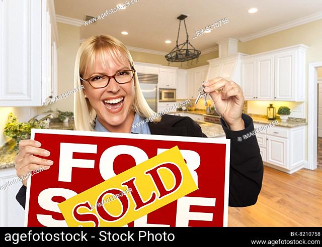 Happy young woman holding sold for sale real estate sign and keys inside beautiful custom kitchen