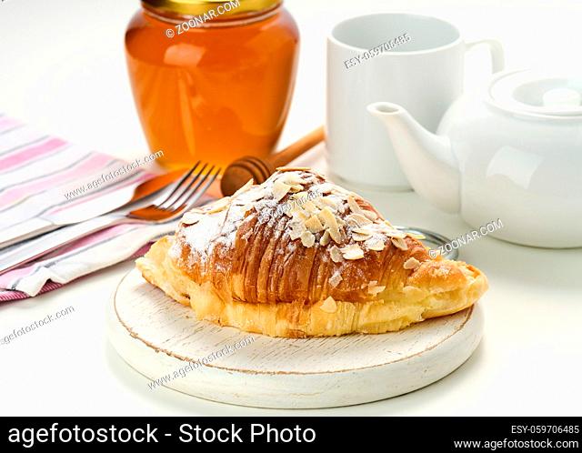 Baked crisp croissant is sprinkled with sugar powder and almond flakes on a wooden board, white ceramic brew and a cup on a white table. Breakfast