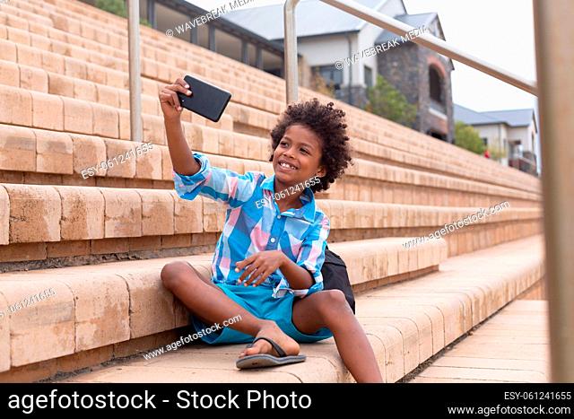 Smiling african american elementary schoolboy taking selfie while sitting on school building steps