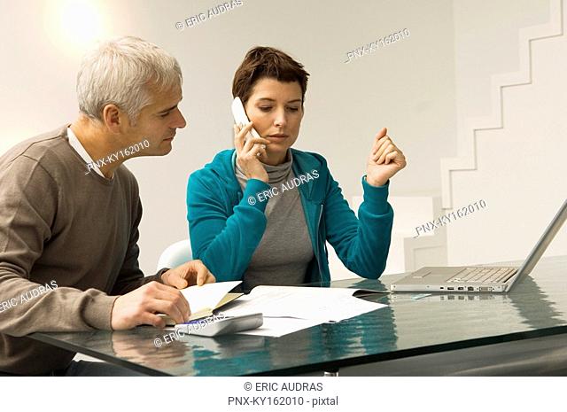 Mature man calculating bills and a mid adult woman talking on a mobile phone beside him