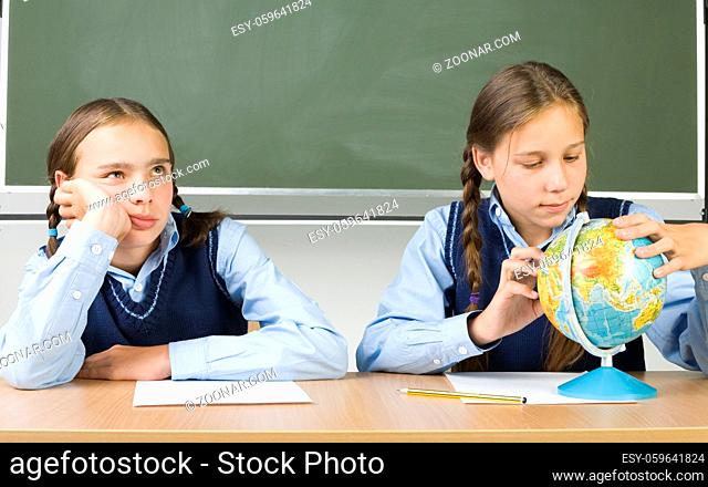 Two girls sitting at desk. On looking bored. Other is searching something on globe. Front view