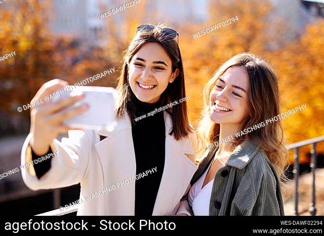 Teenage girl taking selfie with friend through smart phone in autumn park