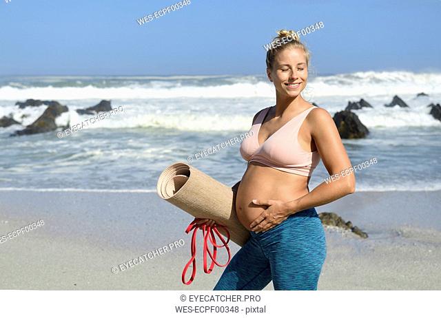 Active pregnant woman holding gym mat on the beach
