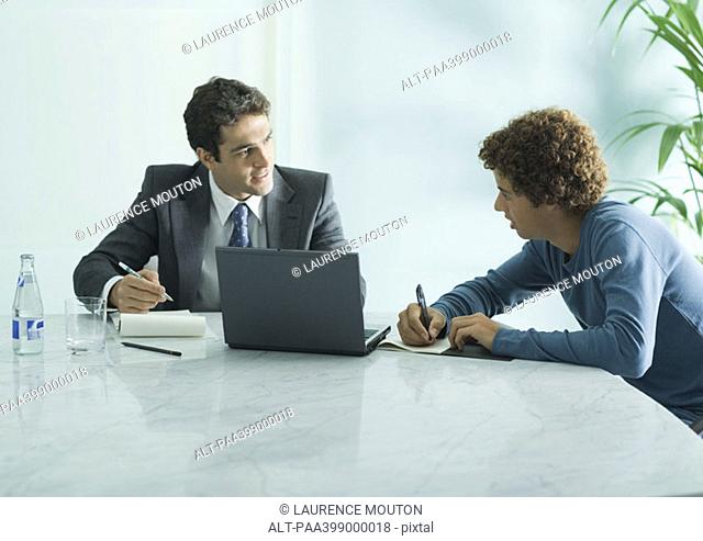 Businessman speaking to casually dressed young adult male, sitting at table