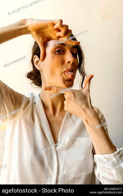 Playful woman sticking out tongue while making finger frame at home