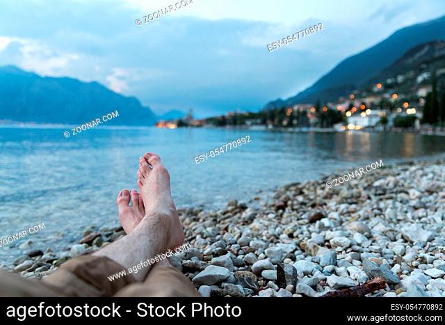 Man is sitting on the pebble beach, enjoying the view over the lake Lago di Garda, evening
