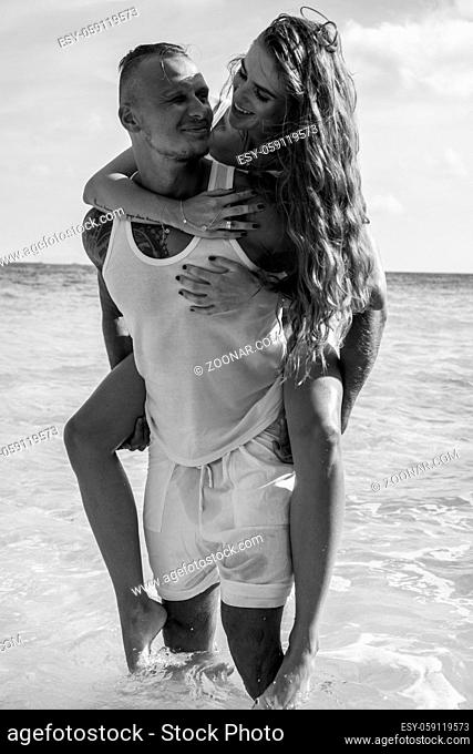 Cheerful handsome man carrying his beautiful girlfriend on his back on the beach over sea and sky background - black and white photo